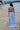 Girl in a light blue dress standing on a beach with water in the background